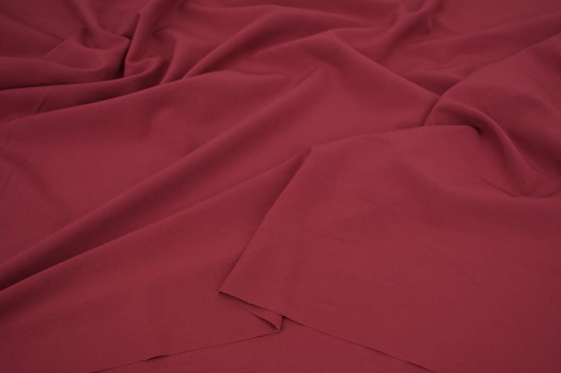May include: A solid dark red fabric with a smooth, matte texture. The material is draped in soft, flowing folds, creating deep shadows and highlights across the surface. The fabric appears opaque and consistent in its deep burgundy red color.