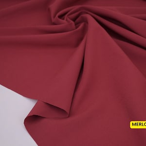 May include: A piece of fabric in a deep, solid merlot red color. The material is draped to create soft, flowing folds and ripples across the surface. A small yellow rectangular label in the bottom right corner displays the text MERLOT.