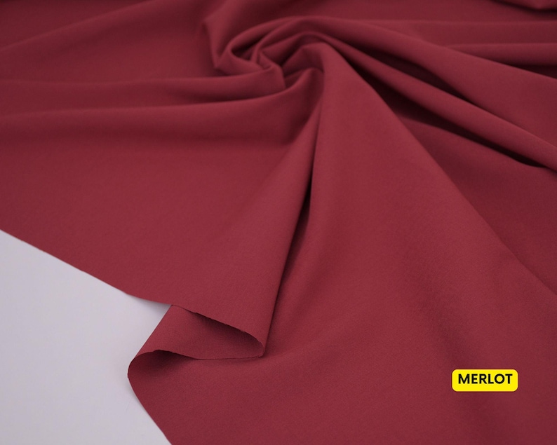 May include: A piece of fabric in a deep, solid merlot red color. The material is draped to create soft, flowing folds and ripples across the surface. A small yellow rectangular label in the bottom right corner displays the text MERLOT.