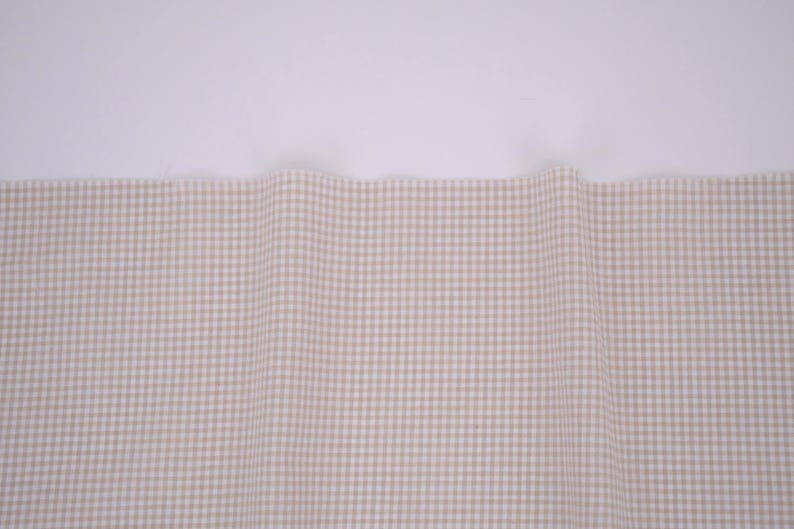 May include: A piece of fabric featuring a small-scale beige and white gingham check pattern. The material is draped to create soft, vertical folds across the surface, displaying a consistent grid design throughout the visible textile area.