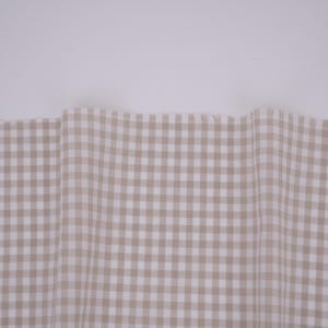 May include: A piece of fabric featuring a light beige and white gingham check pattern. The material is draped in soft, vertical folds, displaying a consistent grid design across the surface with a clean, straight top edge against a plain background.