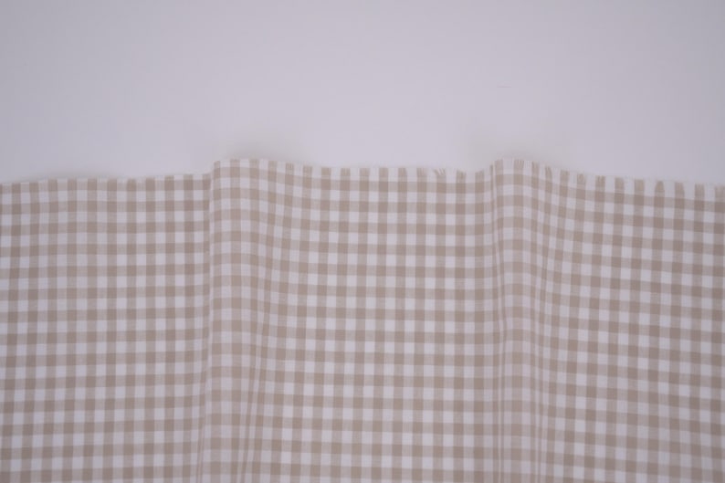 May include: A piece of fabric featuring a light beige and white gingham check pattern. The material is draped in soft, vertical folds, displaying a consistent grid design across the surface with a clean, straight top edge against a plain background.