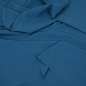 May include: A solid, deep blue fabric with a smooth, matte texture. The material is draped in soft, flowing folds, creating natural shadows and highlights across the surface that emphasize the consistent, uniform color throughout the textile.