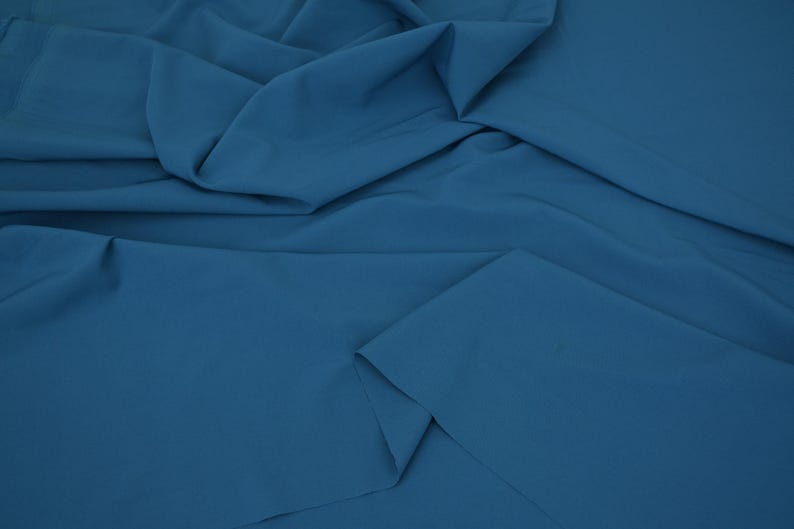 May include: A solid, deep blue fabric with a smooth, matte texture. The material is draped in soft, flowing folds, creating natural shadows and highlights across the surface that emphasize the consistent, uniform color throughout the textile.
