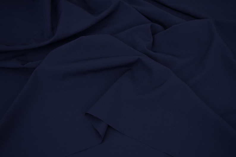 May include: A solid dark navy blue fabric with a smooth, matte texture. The material is draped in soft, flowing folds, creating deep shadows and highlights across the surface that emphasize the consistent, deep blue color of the textile.