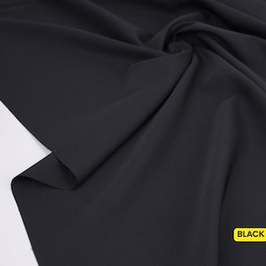 May include: A solid black fabric with a smooth, matte texture, draped in soft folds to create natural shadows and highlights across the surface. A small yellow rectangular label with the word BLACK printed in bold black text is visible.