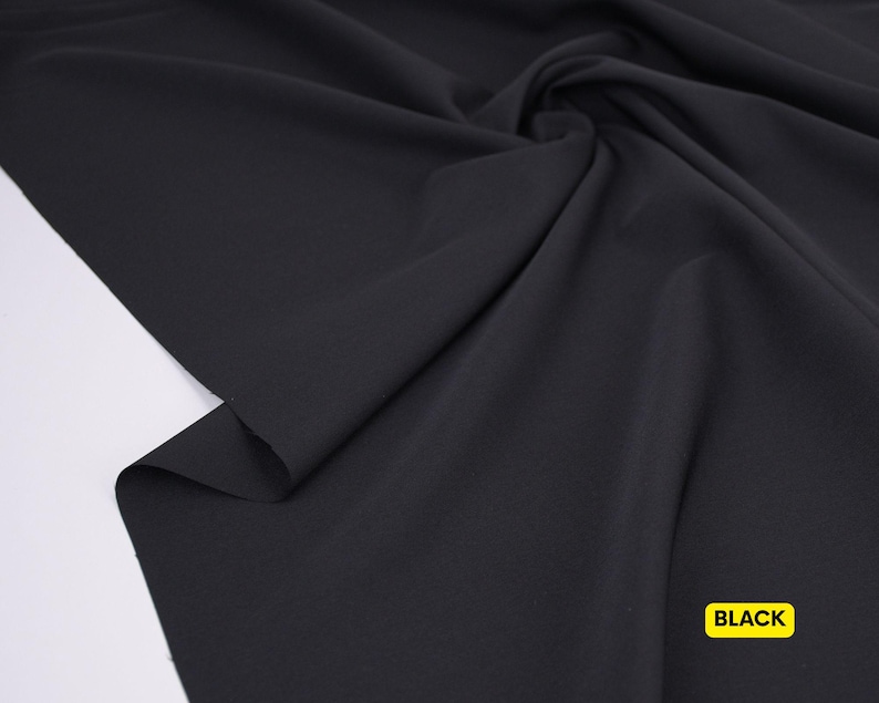 May include: A solid black fabric with a smooth, matte texture, draped in soft folds to create natural shadows and highlights across the surface. A small yellow rectangular label with the word BLACK printed in bold black text is visible.