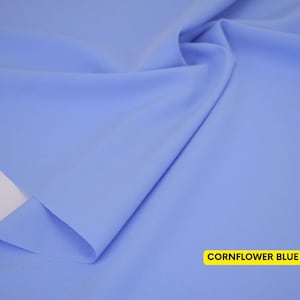 May include: A piece of solid cornflower blue fabric with a smooth, matte texture. The material is draped to create soft folds and shadows across the surface. A rectangular yellow label in the bottom right corner displays the text CORNFLOWER BLUE.