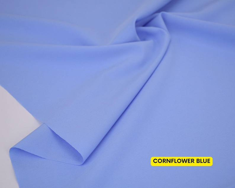 May include: A piece of solid cornflower blue fabric with a smooth, matte texture. The material is draped to create soft folds and shadows across the surface. A rectangular yellow label in the bottom right corner displays the text CORNFLOWER BLUE.