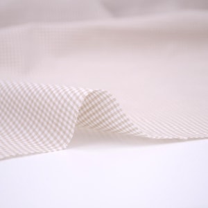 May include: A piece of fabric featuring a small-scale beige and white gingham check pattern. The textile is draped to show soft folds and a smooth texture, highlighting the repeating grid design across the surface of the material.
