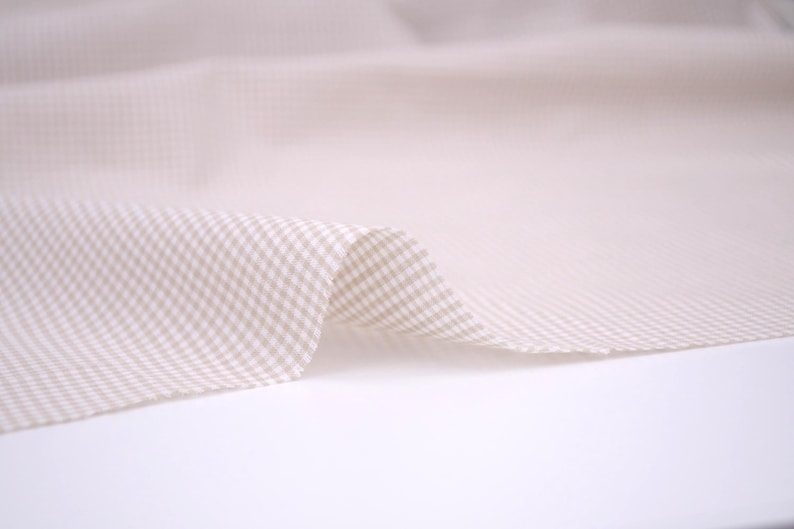 May include: A piece of fabric featuring a small-scale beige and white gingham check pattern. The textile is draped to show soft folds and a smooth texture, highlighting the repeating grid design across the surface of the material.