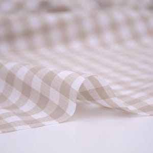 May include: A lightweight fabric featuring a classic gingham pattern in soft beige and white. The material is draped in gentle folds, showcasing the repeating checkered design across the surface with a consistent color palette and texture.