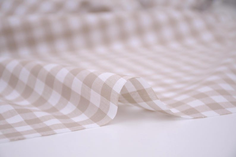 May include: A lightweight fabric featuring a classic gingham pattern in soft beige and white. The material is draped in gentle folds, showcasing the repeating checkered design across the surface with a consistent color palette and texture.