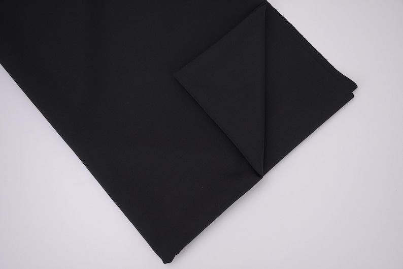 May include: A solid black fabric piece folded at one corner to reveal the texture and depth of the material. The textile features a smooth, matte finish with clean, straight edges and a consistent, deep black color throughout the entire surface.
