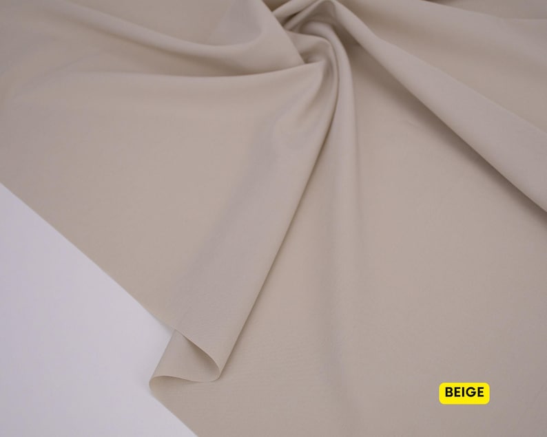 May include: A piece of solid beige fabric draped to create soft folds and curves. The material has a smooth, matte surface texture. A small yellow rectangular label in the bottom right corner contains the black text BEIGE.