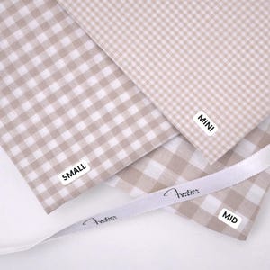 May include: Three swatches of beige and white gingham fabric with white labels reading MINI, SMALL, and MID. A white ribbon with the printed text "Frontier Fabrics" is draped across the fabric pieces, which feature varying check sizes.
