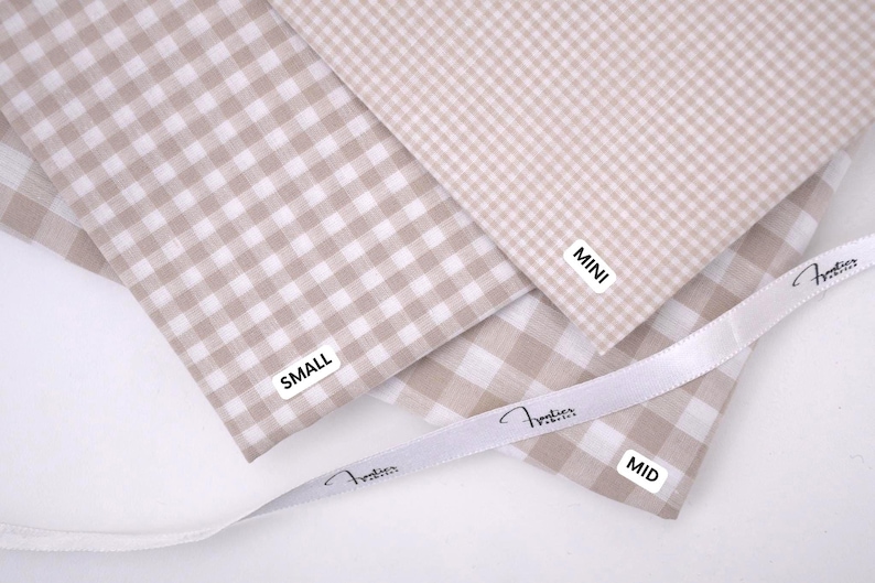 May include: Three swatches of beige and white gingham fabric with white labels reading MINI, SMALL, and MID. A white ribbon with the printed text "Frontier Fabrics" is draped across the fabric pieces, which feature varying check sizes.