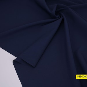 May include: A solid dark navy blue fabric draped in soft folds, featuring a smooth, matte texture. In the bottom right corner, a small yellow rectangular label is clearly visible with the word INDIGO printed in bold black capital letters.