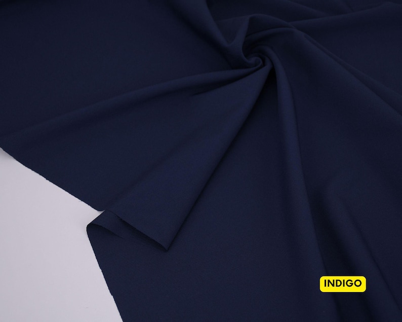 May include: A solid dark navy blue fabric draped in soft folds, featuring a smooth, matte texture. In the bottom right corner, a small yellow rectangular label is clearly visible with the word INDIGO printed in bold black capital letters.