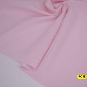 May include: A solid light pink fabric with a smooth texture and soft folds. The material is draped to show its flexibility and drape. In the bottom right corner, there is a small yellow rectangular label with the word ROSE printed in black.