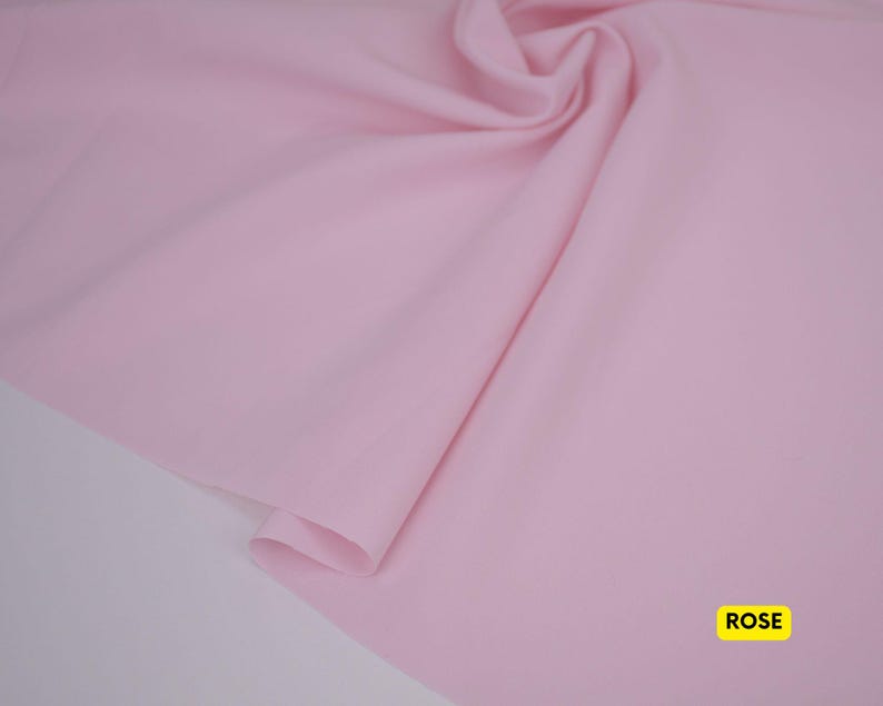 May include: A solid light pink fabric with a smooth texture and soft folds. The material is draped to show its flexibility and drape. In the bottom right corner, there is a small yellow rectangular label with the word ROSE printed in black.