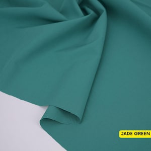 May include: A piece of solid jade green fabric draped to create soft folds and curves. The material has a smooth, matte texture and a consistent deep green color throughout. A small yellow rectangular label in the corner displays the text JADE GREEN.