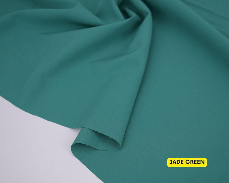 May include: A piece of solid jade green fabric draped to create soft folds and curves. The material has a smooth, matte texture and a consistent deep green color throughout. A small yellow rectangular label in the corner displays the text JADE GREEN.