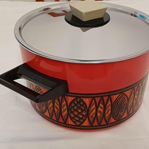 May include: A red cooking pot with a silver lid and black handles. The pot features a decorative band of orange, brown, and black geometric patterns around the base. The lid has a beige square handle.
