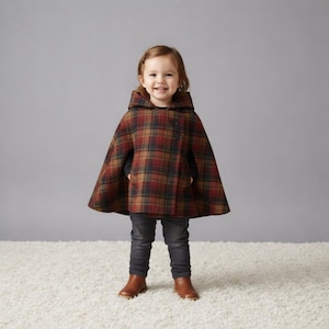 Puede incluir: Un niño lleva una capa con capucha con un estampado de cuadros rojos, marrones y dorados. La capa tiene un cierre de botón. El niño lleva vaqueros grises oscuros y botas marrones. El niño sonríe.