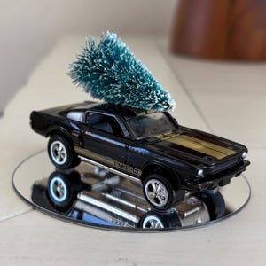 May include: A black toy car with gold stripes and a small green Christmas tree on top. The car is displayed on a small, round mirror. The car is a classic muscle car style.