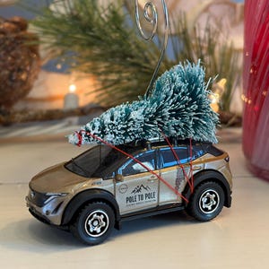 May include: A miniature gold SUV ornament with a small green Christmas tree secured to the roof with red string. The ornament has black wheels and the text "POLE TO POLE" on the side. A silver wire loop is attached for hanging.
