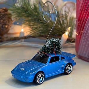May include: A blue toy car with a miniature Christmas tree attached to its roof. The car has black wheels and a silver wire loop for hanging. The ornament is set against a festive background with greenery and a red glass.
