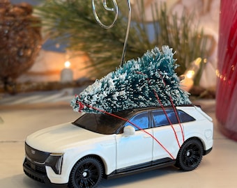 Op de afbeelding: Een witte SUV kerstversiering met een kleine groene kerstboom vastgebonden op het dak met rood touw. De auto heeft zwarte wielen en een zilveren haak. De achtergrond is wazig.