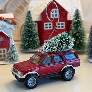 Puede incluir: Un vehículo SUV de juguete en miniatura de color rojo oscuro con un pequeño árbol de Navidad artificial verde sujeto al techo con un hilo rojo fino. Un soporte de alambre metálico plateado en espiral se extiende desde el techo.