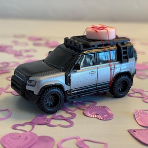 May include: A silver and black toy SUV with a roof rack, a small pink box tied to the roof, and black wheels. The toy is surrounded by pink heart-shaped confetti. The word "Defender" is visible on the hood.