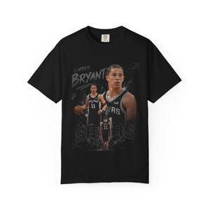 May include: Black t-shirt featuring a basketball player graphic. The design includes the name "Carter Bryant" and the word "Spurs". The player is wearing a jersey with the number 11. The shirt has a crew neck and short sleeves.