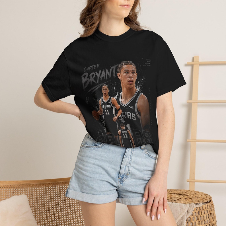 May include: Black t-shirt featuring a graphic of basketball player Carter Bryant in a San Antonio Spurs jersey. The design includes the player's name and jersey number, with additional images of the player. The shirt is a classic crew neck style.