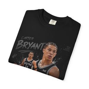 May include: Black t-shirt featuring a graphic of basketball player Carter Bryant. The design includes his name in a stylized font and a portrait of him in a basketball uniform. The shirt also has the words "Defense, Energy, Potential, Squad Future."