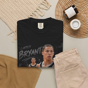 May include: Black t-shirt featuring a portrait of Carter Bryant with the name in a distressed font. The shirt includes a small image of Bryant and the Comfort Colors logo. Beige pants and a woven mat are also visible.