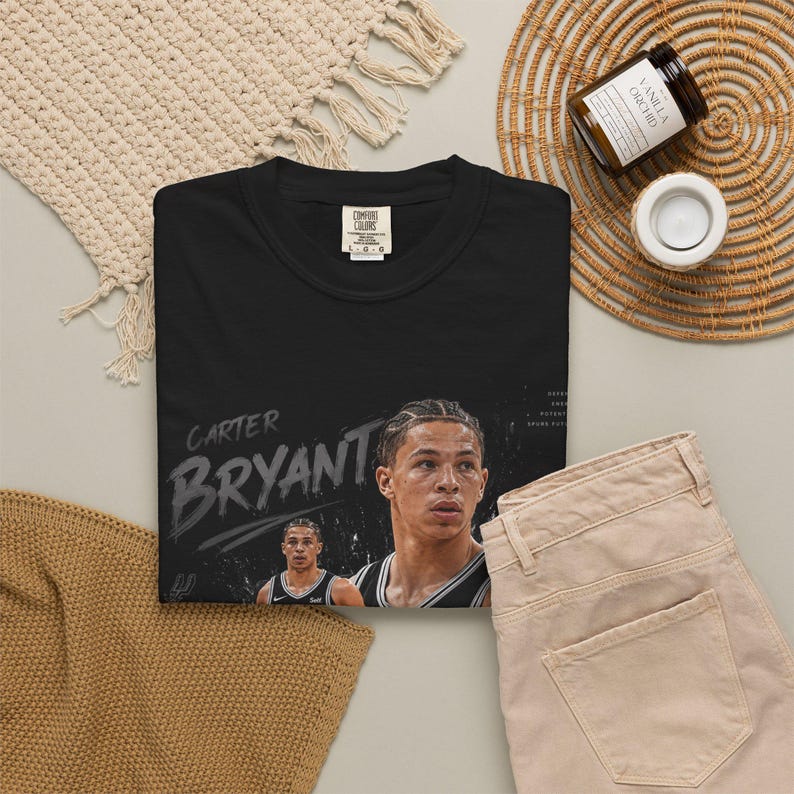 May include: Black t-shirt featuring a portrait of Carter Bryant with the name in a distressed font. The shirt includes a small image of Bryant and the Comfort Colors logo. Beige pants and a woven mat are also visible.
