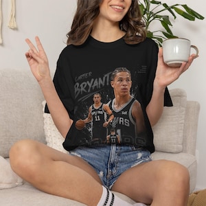 May include: Black t-shirt featuring a graphic of a basketball player with the name "Carter Bryant" in white. The player is wearing a black and silver jersey with the number 11. The shirt has a crew neck and short sleeves.