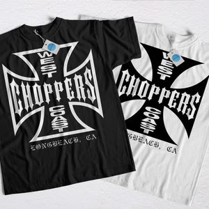 Puede incluir: Dos camisetas, una negra y otra blanca, con un gran diseño de cruz y las palabras "West Coast Choppers" en fuente gótica. El texto "Long Beach, CA" está debajo. Las camisetas tienen etiquetas.