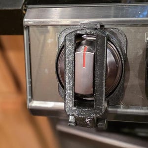 Puede incluir: Primer plano de una cubierta de seguridad para perilla de estufa negra instalada en una estufa de acero inoxidable. La cubierta es de plástico negro y tiene forma rectangular. La perilla de la estufa es plateada con un indicador rojo.