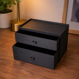 May include: A black desktop storage organizer featuring two stacked drawers. Each drawer is equipped with a small, semi-circular black pull handle. The unit has a flat top surface and a rectangular frame, constructed with a matte finish throughout.