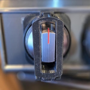 May include: A black plastic stove control knob featuring a central silver metallic insert with a vertical red indicator line. The knob has a rectangular frame design and is mounted on a stainless steel control panel surface.