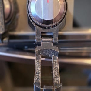 以下が含まれることがあります： 赤色のインジケーターラインが付いた、黒とシルバーの機械部品のクローズアップ。 部品には円形のダイヤルと、テキストが書かれたヒンジ付きの下部セクションがあります。 背景はぼやけています。