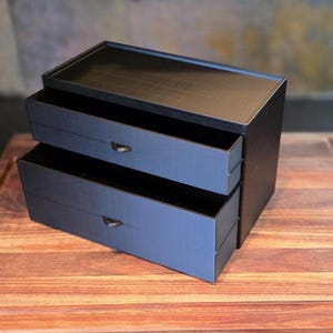 May include: A matte black desktop storage unit featuring two horizontal drawers stacked vertically. Each drawer is equipped with a small, centered black pull tab for opening. The rectangular organizer has a smooth, solid finish and a flat top surface.
