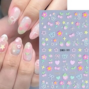 K&ouml;nnte beinhalten: Ein Set Nagelkunst-Aufkleber und eine Hand mit dekorierten N&auml;geln. Die Aufkleber zeigen rosa Blumen, Erdbeeren, Schleifen und Sterne. Die N&auml;gel sind mit einem rosa-wei&szlig;en Farbverlauf bemalt, mit &auml;hnlichen Verzierungen.