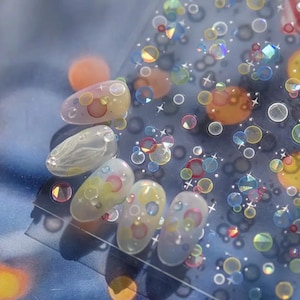 Peut inclure: Cinq faux ongles avec une base transparente, décorés de cercles colorés, d'étoiles et d'accents irisés. Le design comprend des nuances de jaune, de rose, de bleu et d'orange, créant un look ludique et artistique. Les ongles sont disposés sur une surface bleue.