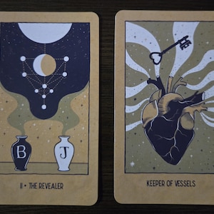 May include: Two tarot cards with beige backgrounds. The first card, "II - The Revealer," features a dark blue and white celestial design above two vases. The second card, "Keeper of Vessels," shows a heart with a key.